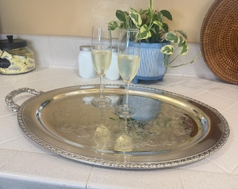 Può includere: Un vassoio ovale d'argento con dettagli ornati contiene due flûte di champagne piene di una bevanda frizzante. Sullo sfondo, un vaso a righe blu e bianche con piante.