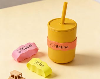 May include: A yellow silicone cup with a matching lid and straw, featuring an orange band printed with the name Belina and a small animal icon. Two additional silicone bands in pink and yellow are printed with the names Claral and John.