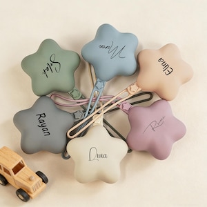 May include: A collection of star-shaped baby pacifier clips in various pastel colors, including green, blue, pink, purple, and beige. Each clip has a name printed on it in a cursive font. A small wooden toy car is in the lower left corner.