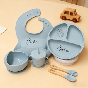 May include: A baby feeding set in light blue, including a bib, divided plate, bowl, sippy cup, and utensils. The set features a raccoon graphic and the name "Cedric". A wooden toy car is also present.