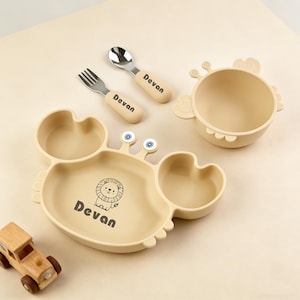 以下が含まれることがあります： ベージュ色のカニ型ベビー食器セット。プレート、ボウル、フォーク、スプーンが含まれています。プレートにはライオンのグラフィックと「Devan」の名前があります。スプーンとフォークの柄にも「Devan」と印刷されています。木製のおもちゃの車もあります。