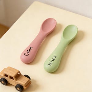 Könnte beinhalten: Zwei Babylöffel, einer rosa und einer grün, mit abgerundeten Schalen und langen Griffen. Auf dem rosa Löffel steht der Name "Calum", auf dem grünen Löffel der Name "Miles". Ein kleines Spielzeugauto aus Holz befindet sich in der linken unteren Ecke.
