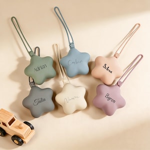 May include: A collection of six star-shaped pacifier clips in various pastel colors: green, blue, peach, gray, cream, and purple. Each clip has a matching strap and is personalized with a name. A small wooden toy car is in the lower left corner.