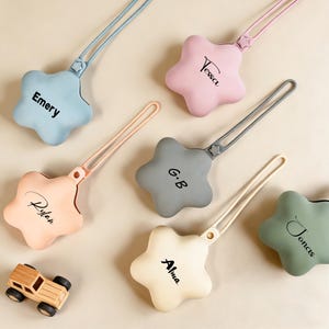 May include: A collection of star-shaped pacifier holders in various pastel colors, each with a name printed on it. The holders have a loop for attaching to a pacifier and a strap. A small wooden toy car is also present.