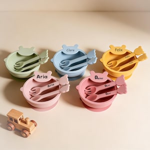 Pode incluir: Conjunto de conjuntos de alimentação para bebés em cores pastel. Cada conjunto inclui uma tigela, um garfo e uma colher. As tigelas são verdes, azuis, amarelas e rosas, com nomes como "Aria" e "Clara". Um comboio de brinquedo de madeira está em primeiro plano.