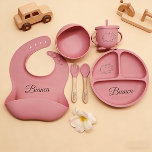 May include: A pink baby feeding set with a bib, bowl, divided plate, cup, and utensils. The set includes a bib with the name "Bianca", a plate and cup with a hippo design, and wooden-handled utensils.