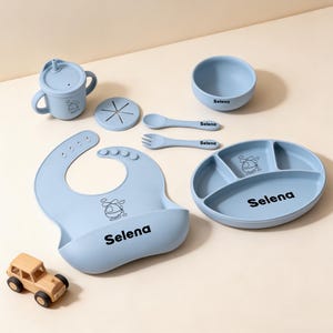 Könnte beinhalten: Ein hellblaues Baby-Ess-Set, bestehend aus Lätzchen, Teller, Schüssel, Becher, Löffel und Gabel. Das Set hat ein Cartoon-Design und den Namen "Selena" auf mehreren Teilen. Ein kleines Holzspielzeugauto ist ebenfalls vorhanden.