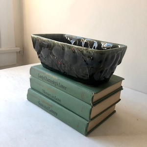 May include: A dark green glazed ceramic planter with a textured surface. The planter is sitting on a stack of three green books. The books are titled "Lady Chatterley's Lover", "Women in Love", and "Sons and Lovers".