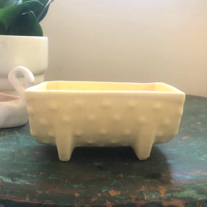 May include: A small, off-white ceramic planter with a textured surface and four short legs. The planter is rectangular in shape and has a slightly rounded bottom.