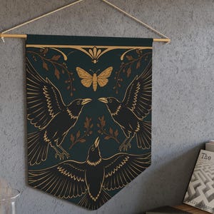 May include: A dark teal fabric wall banner featuring a gold-colored illustration of three black crows with spread wings, a central moth, and leafy branches. The banner is suspended from a wooden dowel with a white cord for hanging on a wall.