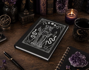 Può includere: Un taccuino nero con un'illustrazione bianca di una dea, serpenti e lune crescenti. Il taccuino è su una superficie di legno con una penna, una candela e altri oggetti a tema occulto.