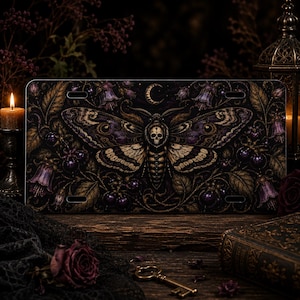 May include: A rectangular metal license plate featuring a central moth with a skull-patterned thorax, surrounded by dark purple bell-shaped flowers, dark foliage, and berries. A crescent moon is positioned above the moth against a dark background.