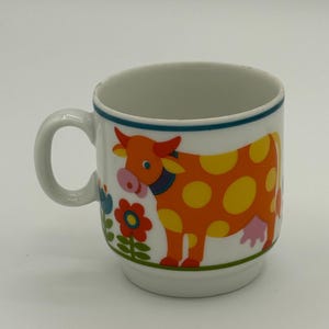May include: White ceramic mug featuring a cartoon cow design. The cow is orange with yellow spots and a pink udder. The mug has a blue rim and a handle. Floral and foliage elements are also present on the mug.