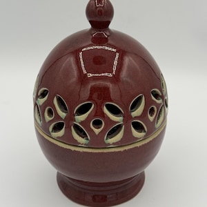 May include: A maroon ceramic incense burner with a lid and a base. The spherical body features floral cutouts, and the lid has a small knob. The base is a slightly lighter shade of maroon. The incense burner is a decorative item.