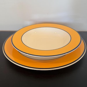Op de afbeelding: Een bord en een kom, op elkaar gestapeld. Beide zijn van keramiek en hebben een oranje buitenkant met een donkerblauwe rand. De binnenkant van de kom is crèmekleurig. Het bord ligt onder de kom.