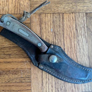 May include: An Old Timer fixed-blade knife with a textured brown handle and a silver metal bolster, secured in a dark brown leather sheath with a metal snap closure and a thin leather lanyard attached to the pommel of the knife handle.