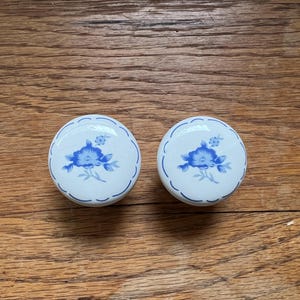 May include: Two round ceramic cabinet knobs with a white base and a blue floral pattern. Each knob features a central blue flower design surrounded by a thin blue scalloped border line that follows the circular edge of the hardware piece.