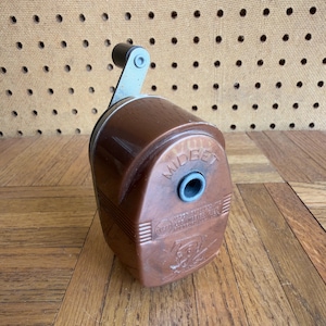 May include: A brown plastic manual pencil sharpener with a dark metal hand crank on top. The front face features the embossed text MIDGET, the APSCO brand logo, and additional manufacturer information. The sharpener has a rounded, oval-shaped body.