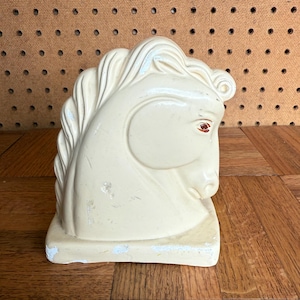 Op de afbeelding: Crèmekleurige keramische paardenhoofdsculptuur. Het paardenhoofd is gebeeldhouwd in een klassieke stijl, met gedetailleerde manen en gelaatstrekken. De sculptuur heeft een rechthoekige basis en is licht beschadigd.