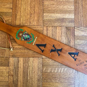 May include: A wooden paddle featuring a leather hanging loop, a colorful crest, and the Greek letters Lambda Chi Alpha in black. The surface includes the engraved text TO KUHN '63 FROM DAVE '65, showcasing a smooth, polished finish on the wood.