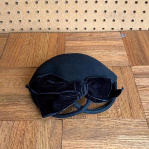 May include: A black pillbox hat with a large velvet bow. The hat has a rounded crown and a structured brim. The bow is a prominent feature, adding a touch of elegance. The hat is likely made of wool or a similar material.