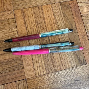 May include: Three pens with different designs are displayed on a wooden surface. One pen has a burgundy barrel, another has a light blue marble design, and the third is bright pink. All have black tips.