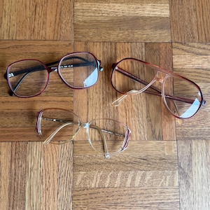 May include: Three pairs of eyeglasses with different frame styles. One pair has a burgundy frame with a rectangular shape. Another pair has a light brown aviator frame. The third pair has a light brown frame with a decorative stripe.