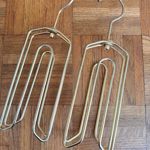 May include: Two gold-toned metal hangers featuring a multi-tiered, open-frame design with a central swivel hook. Each hanger consists of several parallel rectangular loops connected at the top, providing a structured form for organizing garments.