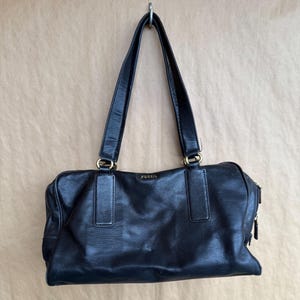 May include: Black leather Fossil handbag with two long handles and gold-colored hardware. The bag has a rectangular shape with a zipper closure and the brand name "Fossil" is visible on the front.