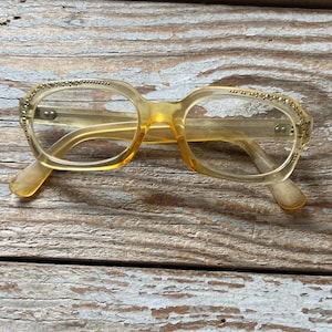 May include: Translucent amber-colored eyeglasses featuring a rectangular frame shape. The upper outer corners of the frames are embellished with small, metallic gold-toned studs arranged in a decorative pattern along the brow line.