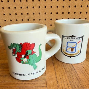 May include: Two white ceramic mugs. The front mug features a green alligator wearing a red shirt with the text TOUGHEST GATOR GOING. The back mug displays a shield-shaped emblem with the text U.S.S. DUBUQUE LPD OUR COUNTRY HERITAGE & FUTURE.