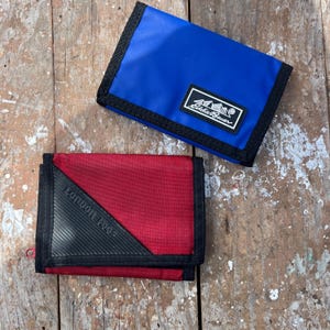 May include: Two wallets, one blue and one red, are displayed on a weathered wooden surface. The blue wallet has a black border and a white Eddie Bauer logo. The red wallet has a black corner with the text "Common Force".