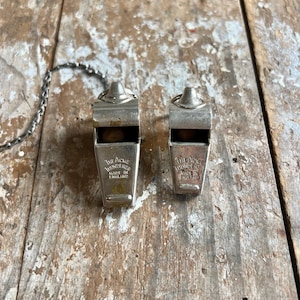 May include: Two vintage metal whistles with the text "The Acme Thunderer Made in England". The whistles are silver-colored and have a small chain attached. They are resting on a weathered wooden surface.