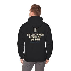 Puede incluir: Una sudadera con capucha negra que presenta texto blanco en la parte posterior que dice THE LOCKED DOOR BETWEEN YOU AND THEM, con cinco líneas verticales sobre el texto y las palabras CORRECTIONS OFFICER impresas en letras azules más pequeñas.