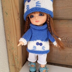 Könnte beinhalten: Eine Puppe trägt eine blaue und weiße gestrickte Katzenmütze, einen blauen Strickpullover mit weißen Streifen und Katzenohren, weiße Hosen und blaue Schuhe. Die Puppe hat gelbes Haar und blaue Augen. Die Puppe steht auf einer weißen Oberfläche mit einem hölzernen Hintergrund.