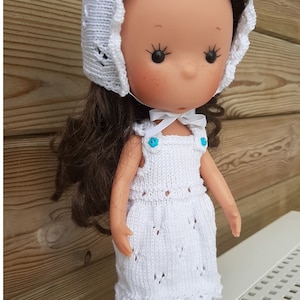 Könnte beinhalten: Ein weißes gestricktes Puppenkleid mit einer weißen Häkelmütze. Die Puppe hat braunes Haar und blaue Augen. Das Kleid hat einen gewellten Saum und eine Schleife am Hals. Die Puppe steht auf einer weißen Oberfläche mit einem Gittermuster.