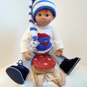 以下が含まれることがあります： 青と白のストライプの帽子をかぶり、赤と青の雪だるまのデザインの白いセーターを着た、青と白のストライプのズボンと青い靴を履いた人形。人形は、ロープが付いた赤と白のウッドスレッドに座っています。