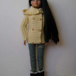 Puede incluir: Una muñeca con un suéter de punto color crema con capucha, jeans azules y botas negras. La muñeca tiene el pelo largo y negro y ojos marrones.