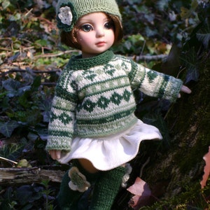 May include: A doll wearing a green and white knitted sweater, a white skirt, and green knitted socks. The doll has a green knitted hat with a white flower on it. The doll is standing in a forest setting.