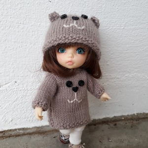 May include: A small doll wearing a brown knitted hat and sweater with a bear face design. The doll has blue eyes and brown hair. The doll is wearing white trousers and brown shoes.