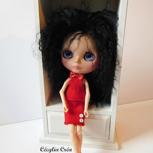 May include: A doll with long black curly hair wearing a red dress with white buttons and white shoes. The doll is standing in a white wooden cabinet with a drawer.