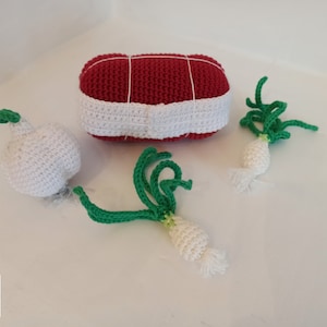 May include: A collection of handmade crochet food items, including a red and white meatloaf, a white garlic bulb, and two green onions. The meatloaf is secured with white string. The items are arranged on a white surface.