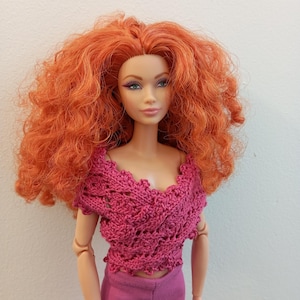 May include: A doll with vibrant red, curly hair, wearing a pink crocheted top and matching pants. The top has a detailed, openwork design. The doll's face features makeup, and it is posed against a plain background.