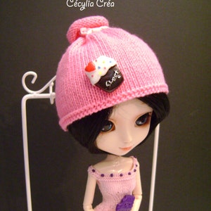 May include: A pink knitted hat with a cupcake button. The hat is on a doll with black hair and blue eyes. The doll is wearing a pink dress with purple accents.