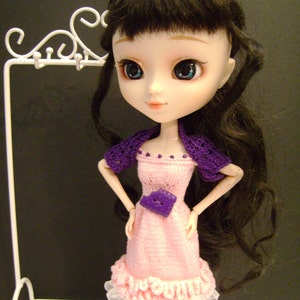 May include: A doll with long black hair wearing a pink knitted dress with a purple crocheted cardigan and a purple button. The doll has large blue eyes and a sweet expression. The doll is standing on a white stand.