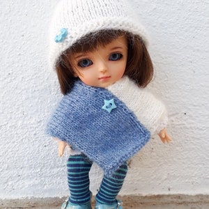 以下が含まれることがあります： 青いポンポンが付いた白いニット帽、青と白のニットポンチョ、青と緑のストライプのレギンス、青い靴を履いた人形。人形は金髪で青い目です。人形は白い表面に立っています。画像の右上に「Cécylia Créa」というテキストがあります。