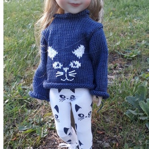 Könnte beinhalten: Eine Puppe trägt einen blauen Strickpullover mit einem weißen Katzenkopfdesign und weiße Leggings mit schwarzen Katzenköpfen. Die Puppe hat blonde Haare und blaue Augen.