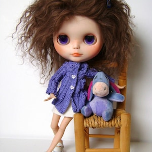 May include: A doll with brown curly hair wearing a purple cardigan and a white skirt. The doll is sitting on a wooden chair with a purple and blue stuffed animal.