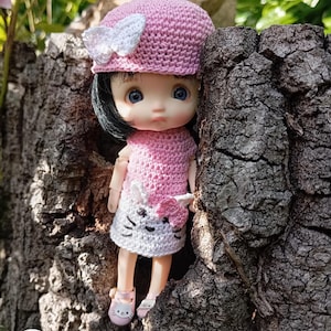 May include: A handmade doll wearing a pink crocheted dress and matching hat with a white bow. The doll has black hair, blue eyes, and pink shoes. The doll is standing next to a tree trunk.