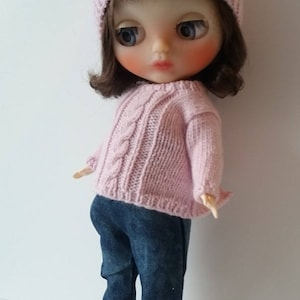May include: A pink knitted doll wearing a pink sweater, blue jeans with plaid trim, and white sneakers. The doll has brown hair and large blue eyes.